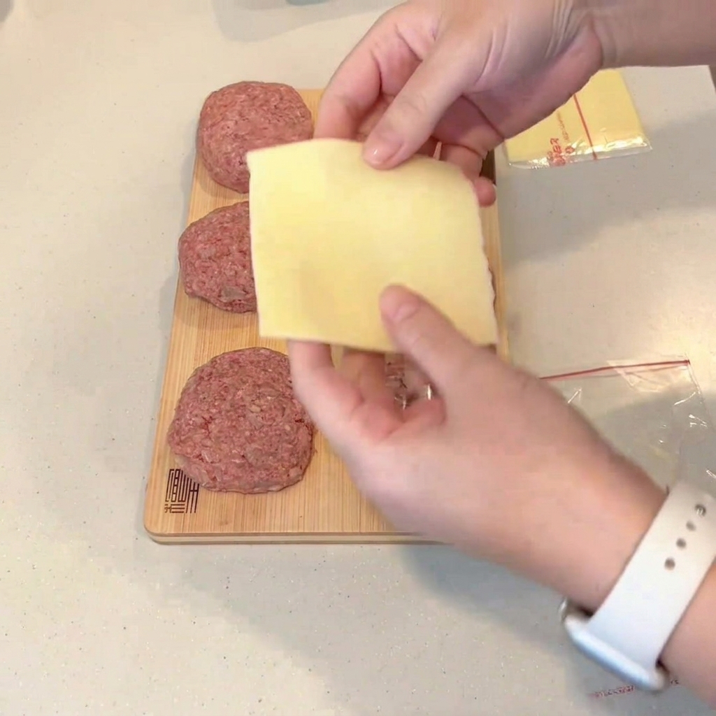 A folded slice of cheese being placed onto the center of a flattened raw meat patty.