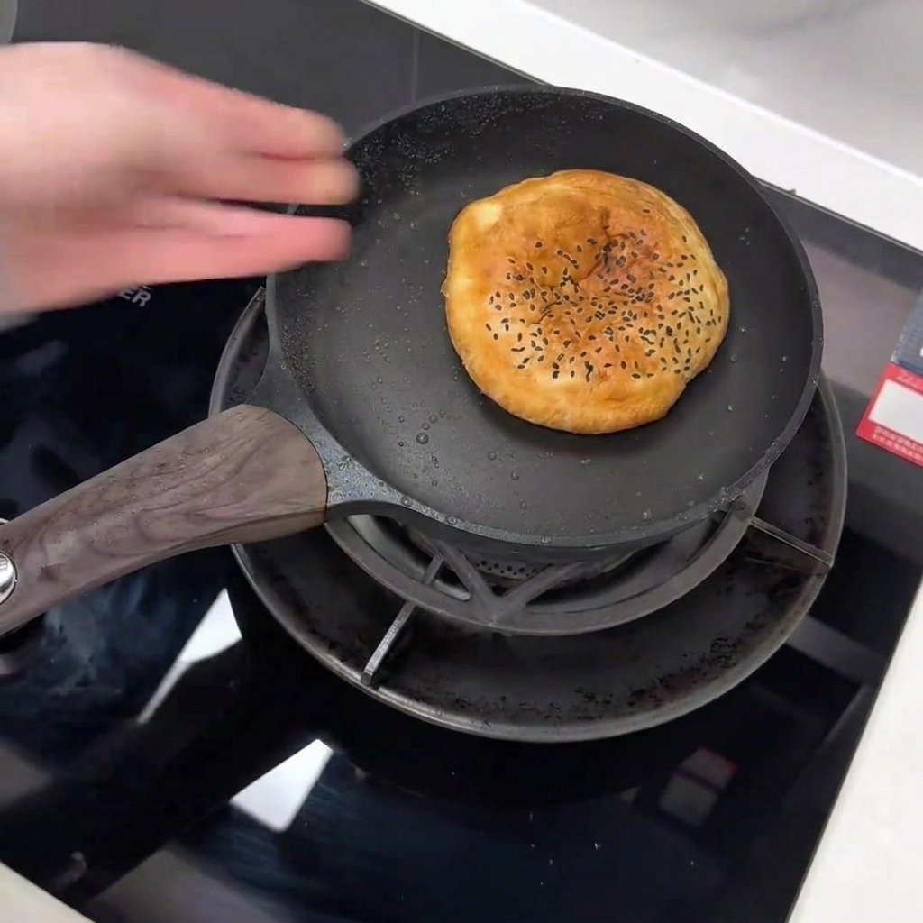 A hand placing a black sesame-topped burger bun into a dark non-stick frying pan.