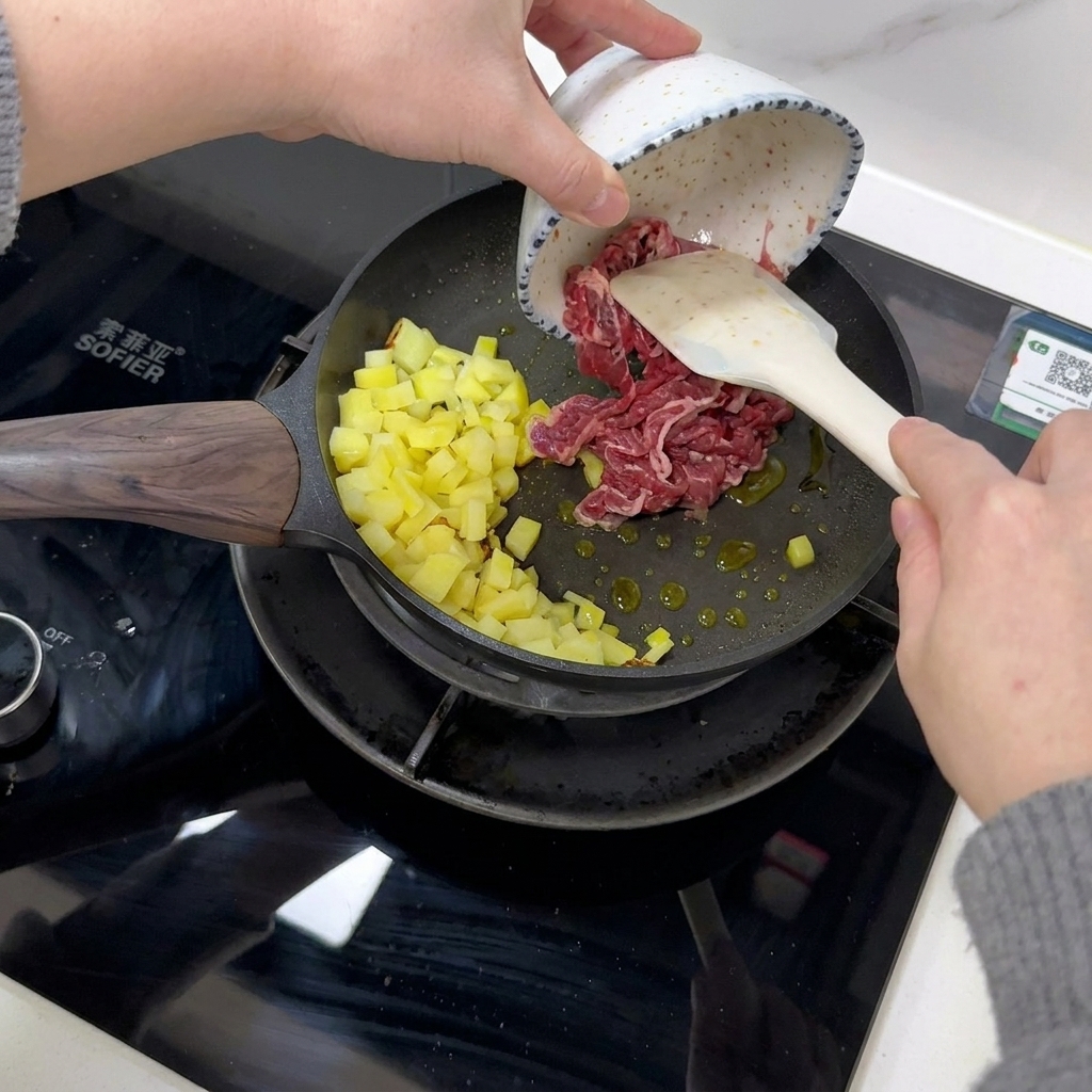 Raw beef slices being added to a pan with cooked diced potatoes using a white spatula.