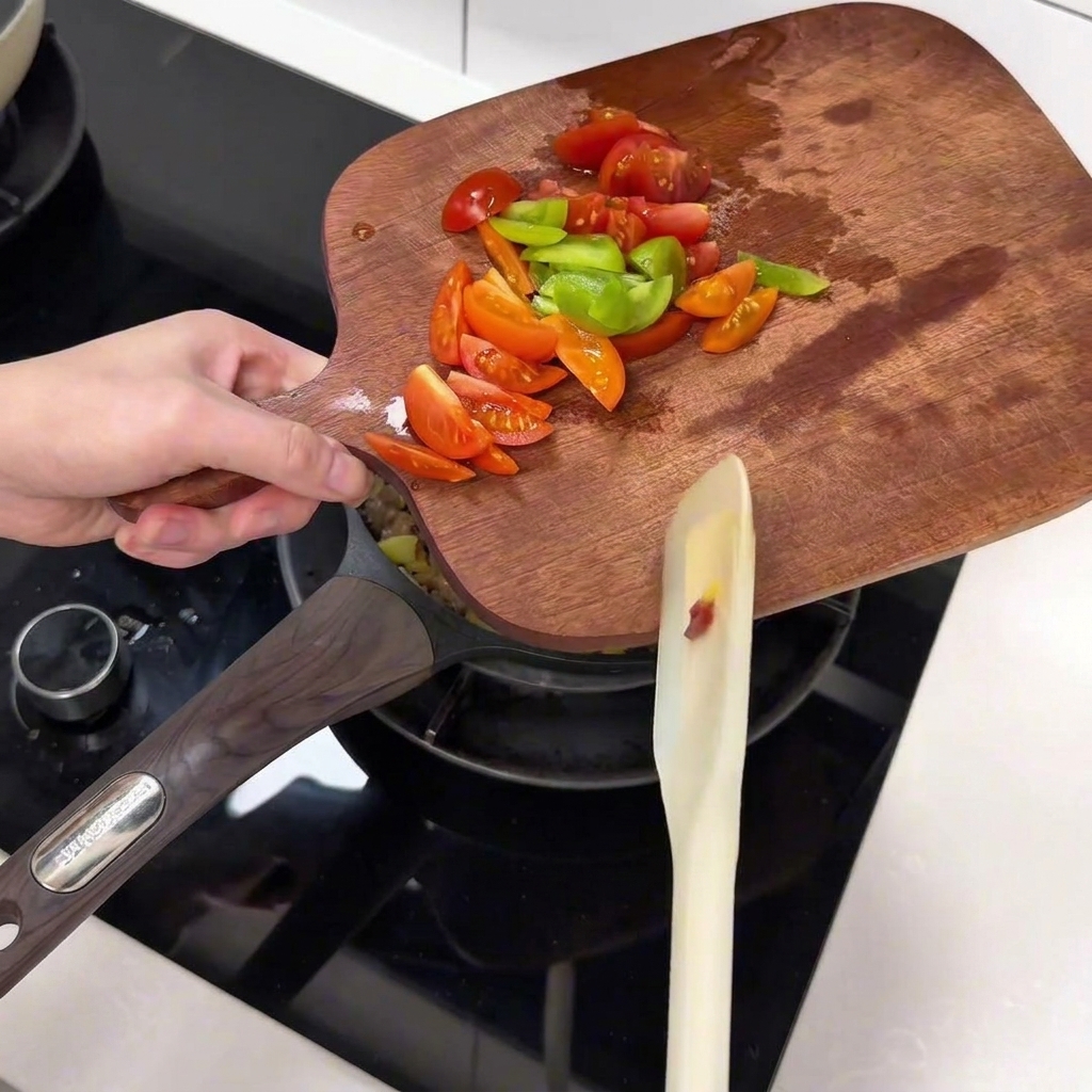 Freshly sliced cherry tomatoes and green pepper slices being transferred from a wooden cutting board into a pan.