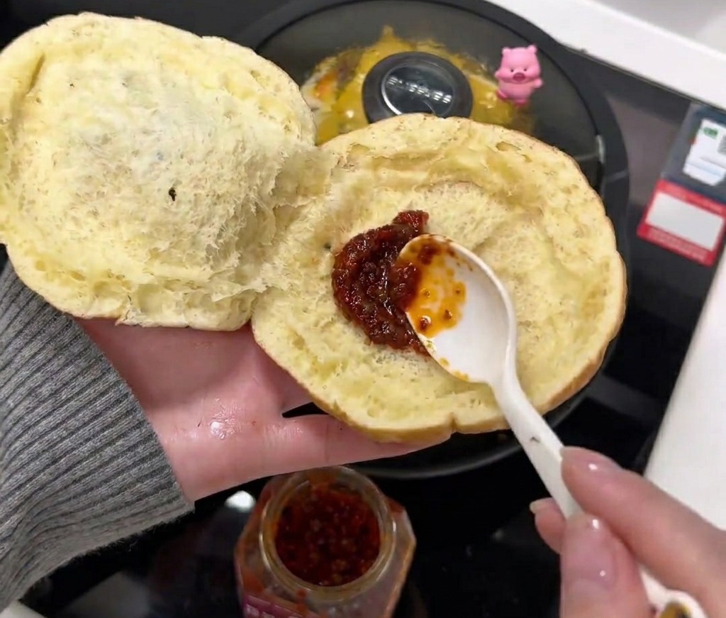 A spoonful of red chili sauce being spread onto the soft inner side of an opened burger bun.