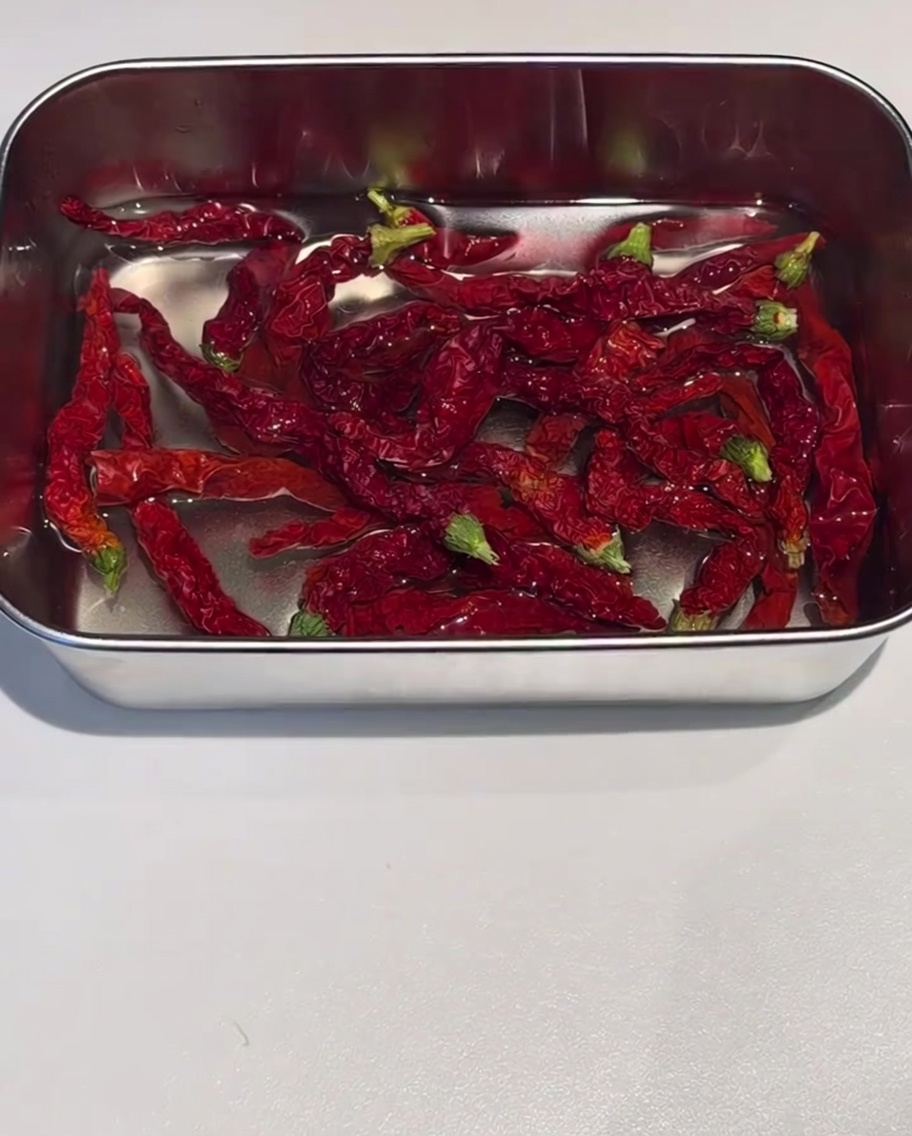Dried red chili skins soaking in a stainless steel container filled with hot water.