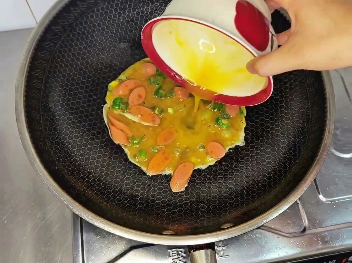 Pouring a mixture of raw beaten eggs, sliced sausage, and diced green chilies from a bowl into a hot wok.