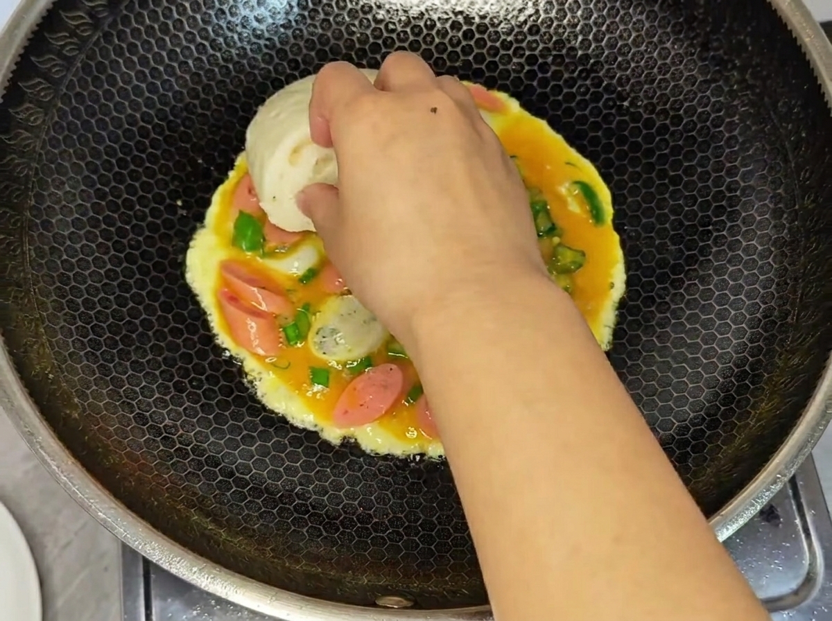 A hand pressing a half of a white steamed bun onto the wet egg and sausage mixture cooking in a wok.