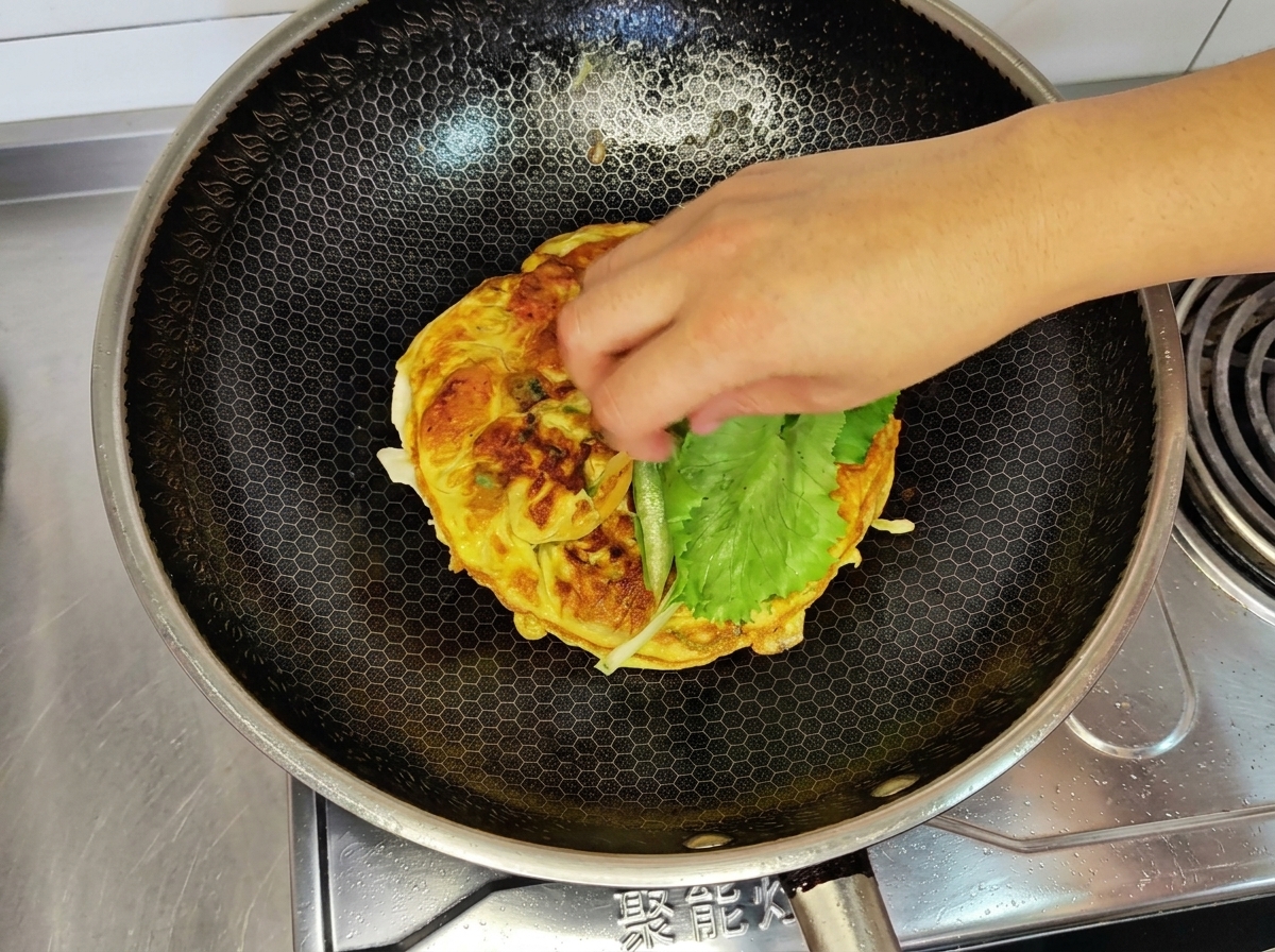 A hand placing a fresh green lettuce leaf on top of the cooked egg omelet in a wok.