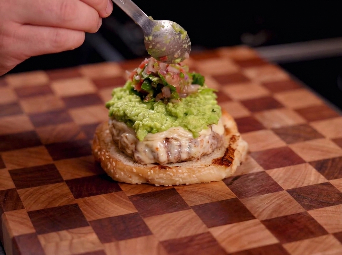 A hand using a spoon to drop fresh red salsa onto a layer of green guacamole on a burger.