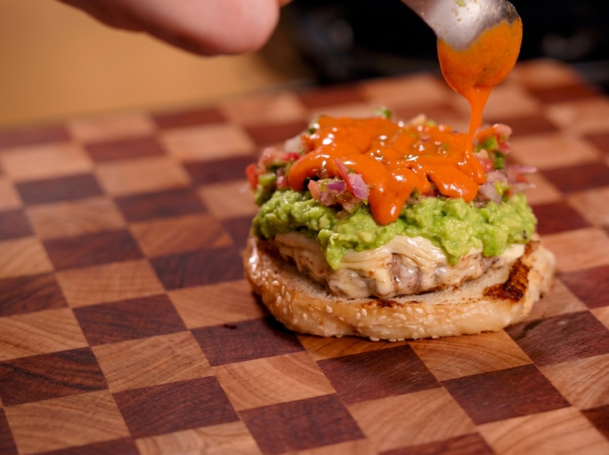 Bright orange hot sauce being drizzled from a spoon over a layer of fresh salsa on a burger.