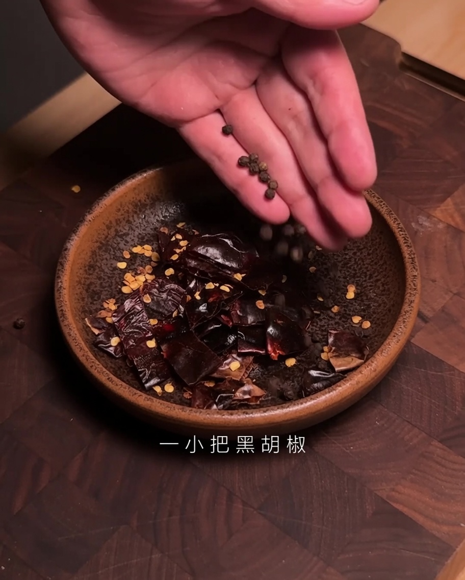 Hands dropping black peppercorns into a bowl containing cut pieces of dried Guajillo chili.
