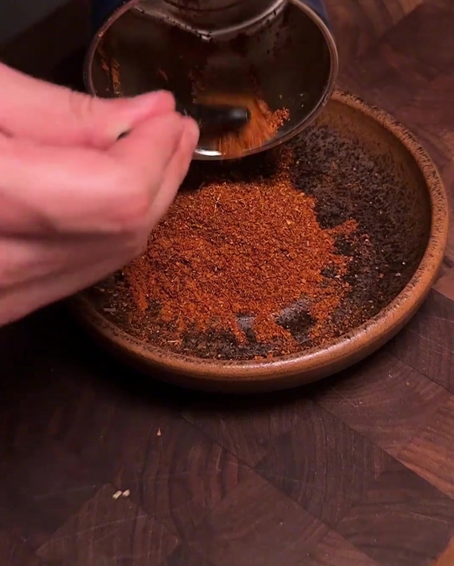 A spice grinder pouring freshly ground, deep red chili and spice powder into a brown ceramic bowl.