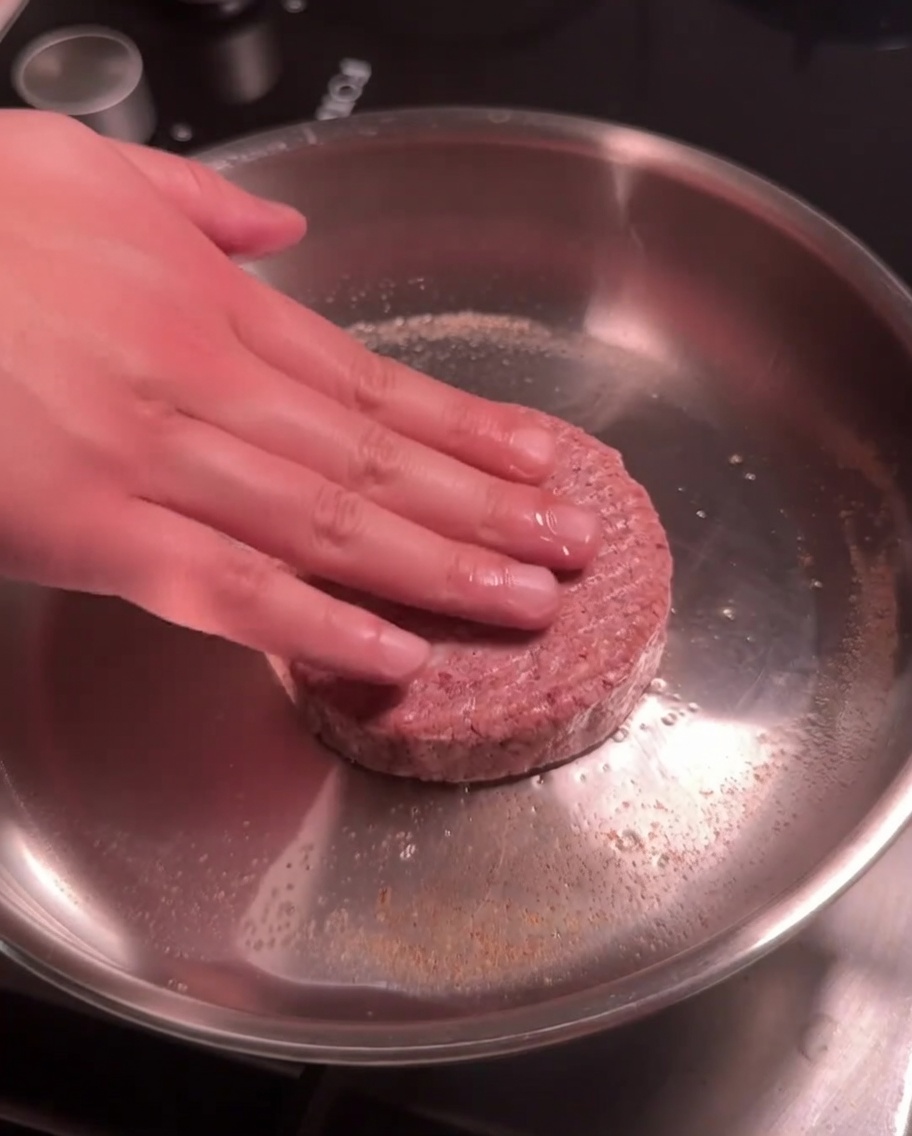 A raw beef patty being pressed down into a hot stainless steel pan for searing.