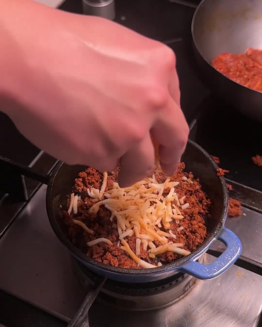 Shredded cheese being sprinkled over the cooked chorizo meat in a small blue pot on the stove.