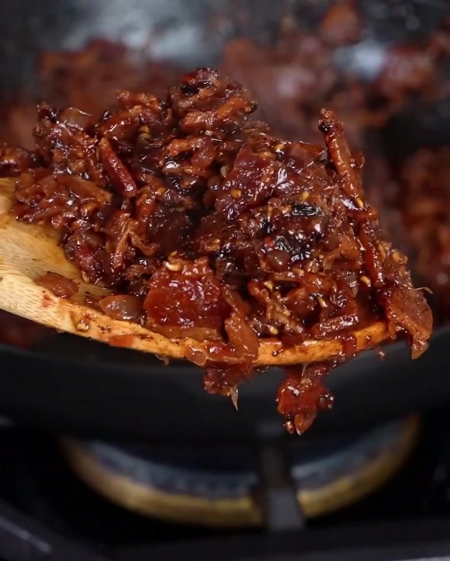 A wooden spoon lifting a scoop of thick, dark reddish-brown homemade bacon jam.