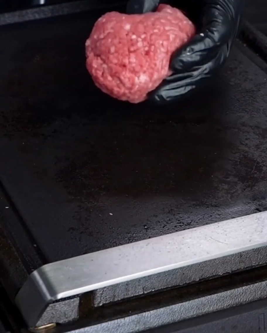 A gloved hand placing a ball of raw 75/25 ground beef onto a black flat top grill.