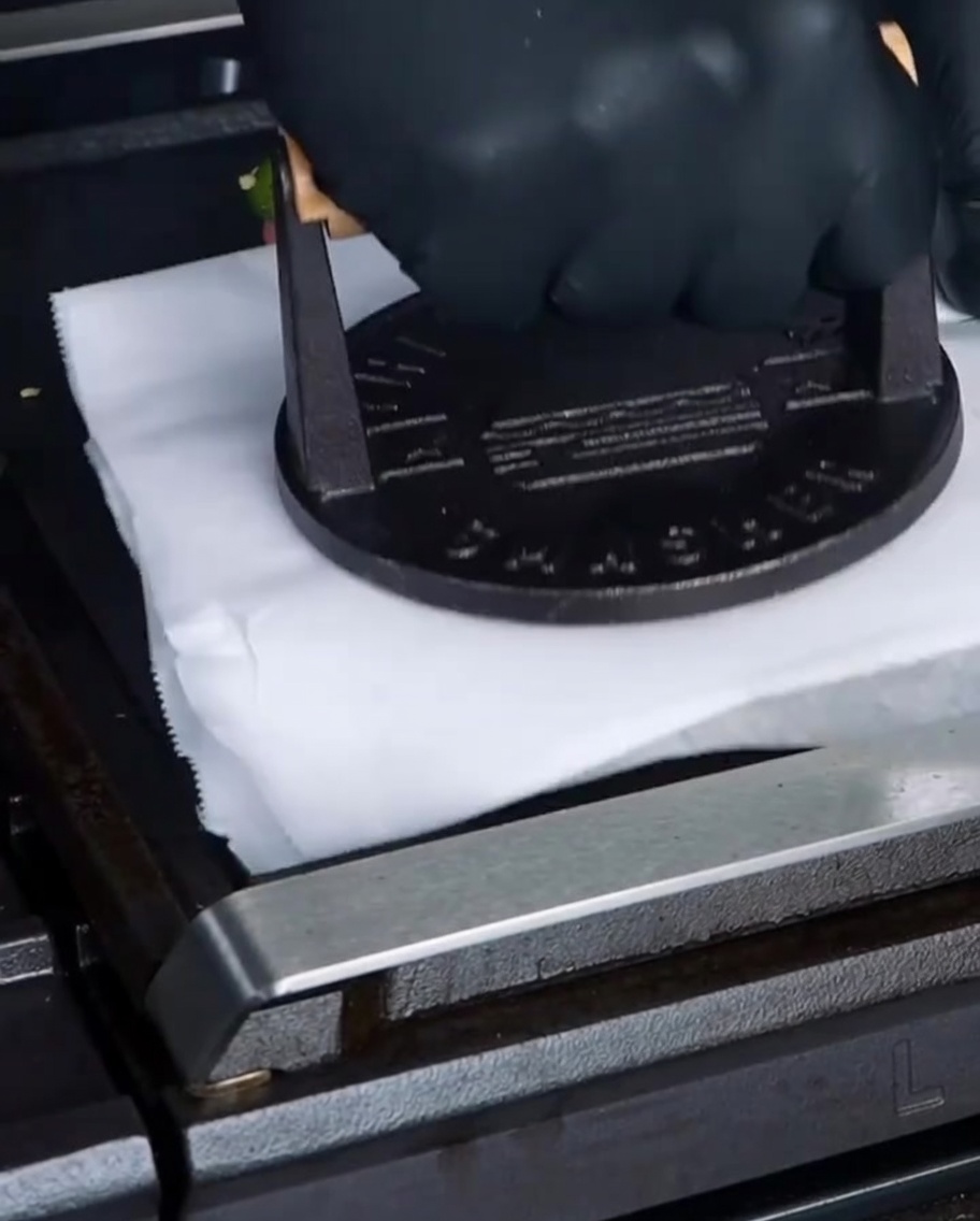 A heavy cast iron burger press pushing down on a piece of parchment paper over a beef patty on the flat top grill.