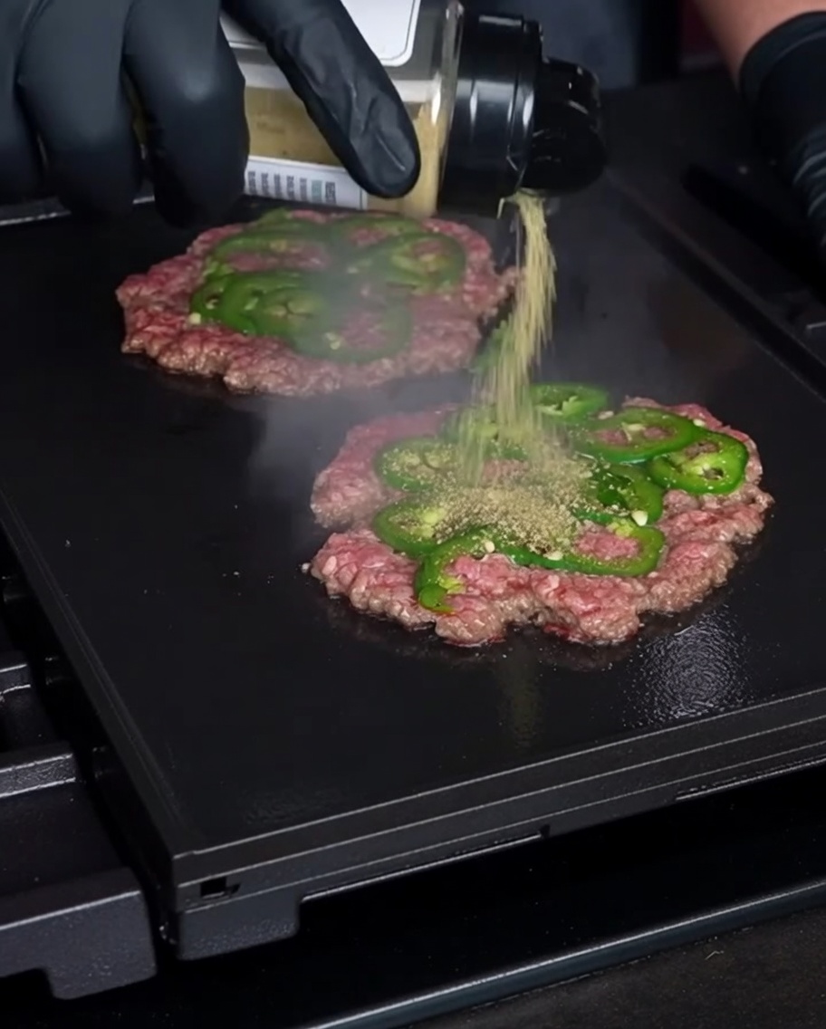 A gloved hand shaking seasoning onto thin, smashed beef patties embedded with fresh jalapeño slices on a hot grill.