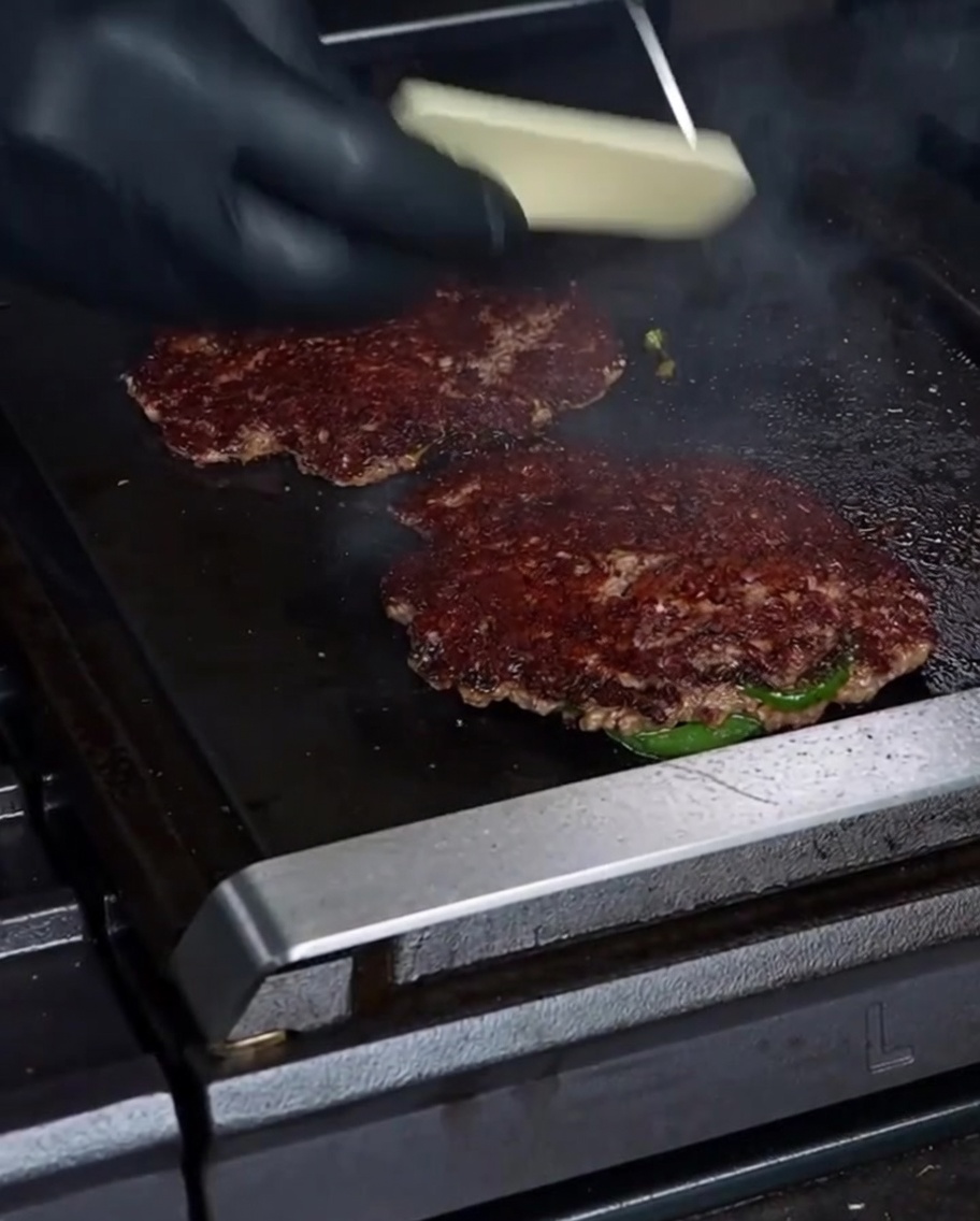 A gloved hand placing a slice of white Havarti cheese onto a beautifully seared smashburger patty on a hot griddle.