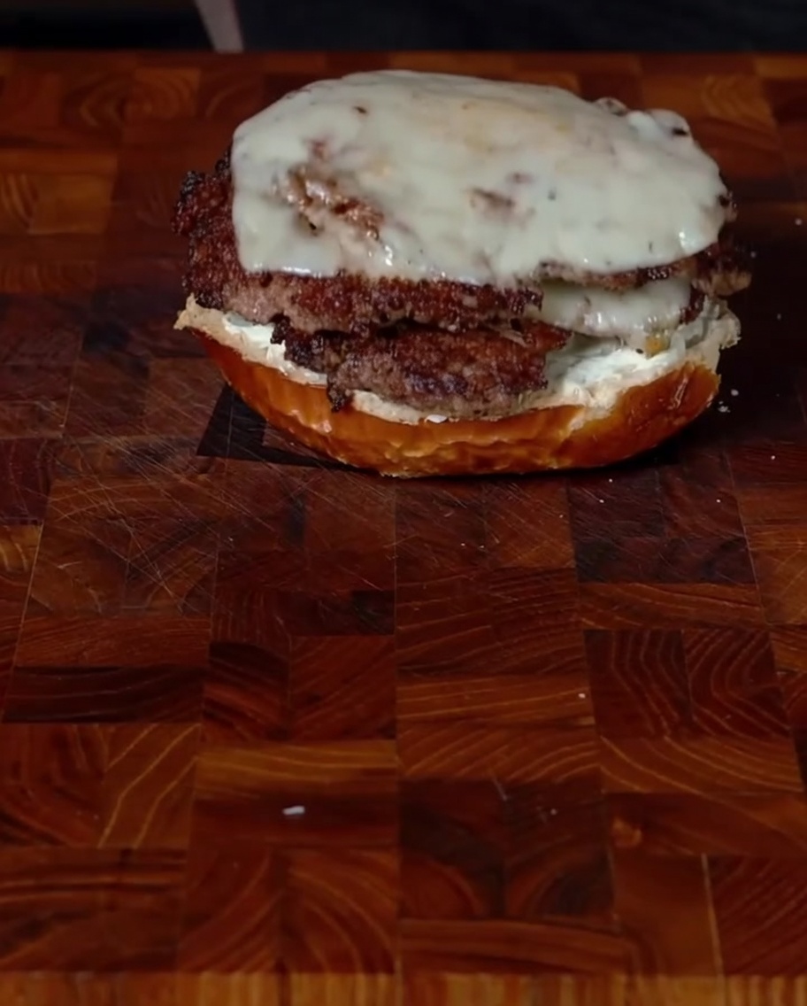 Carefully placing two stacked smashburger patties with jalapeños and melted cheese onto a bottom bun coated with cream cheese.