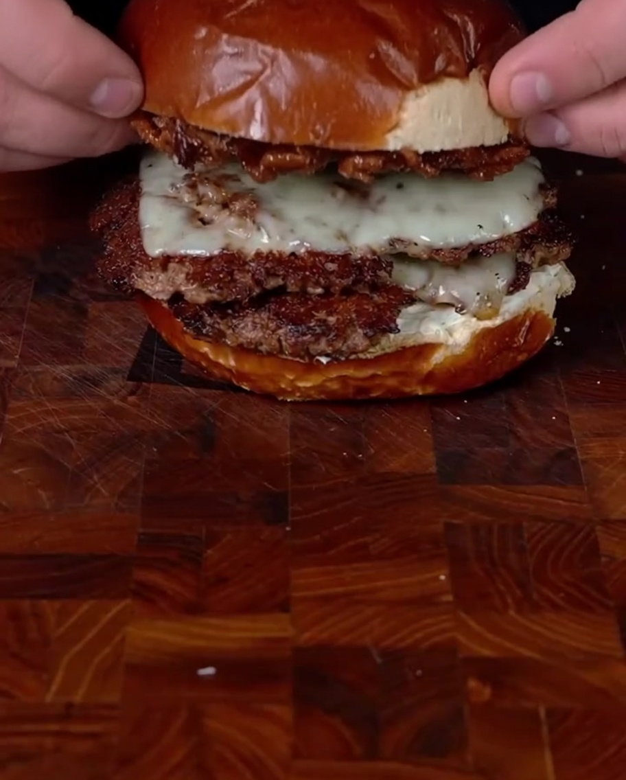 Hands placing the top bun loaded with bacon jam onto the stacked double smashburger to complete the assembly.