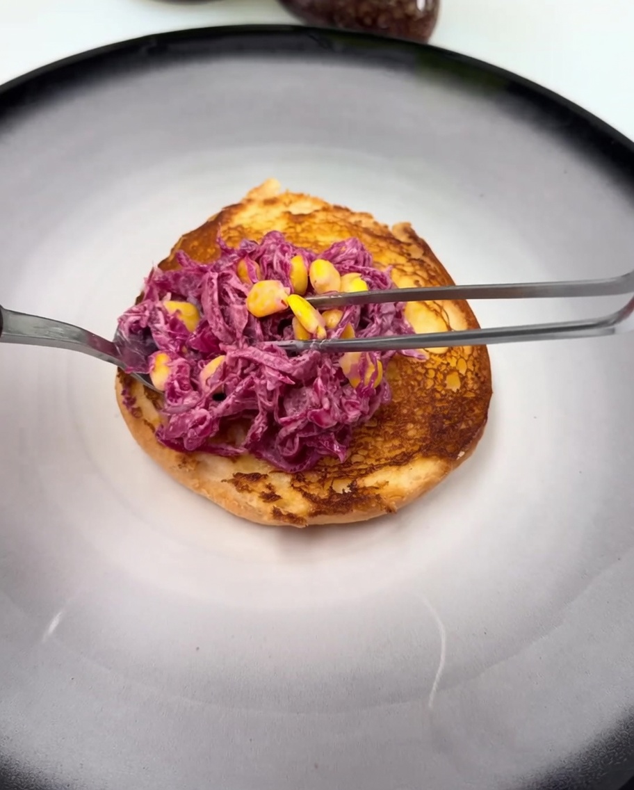 The bottom toasted burger bun topped with a spread of sauce and a portion of the purple cabbage and corn slaw.