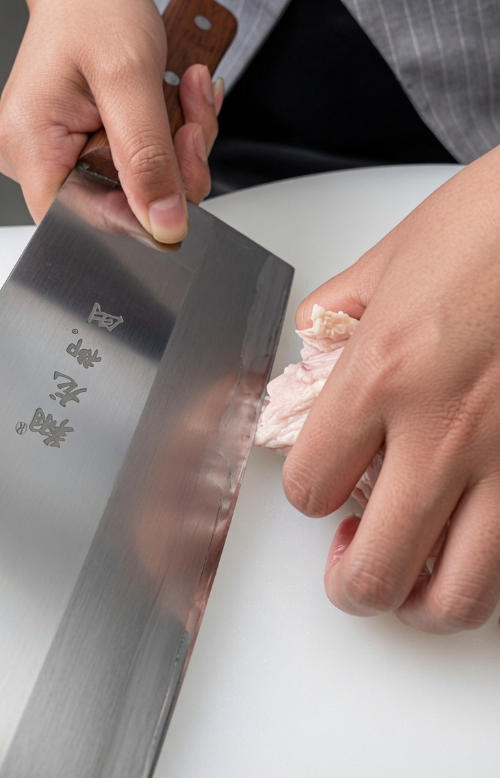 A close-up of a raw chicken leg being deboned on a white surface with a heavy kitchen cleaver.