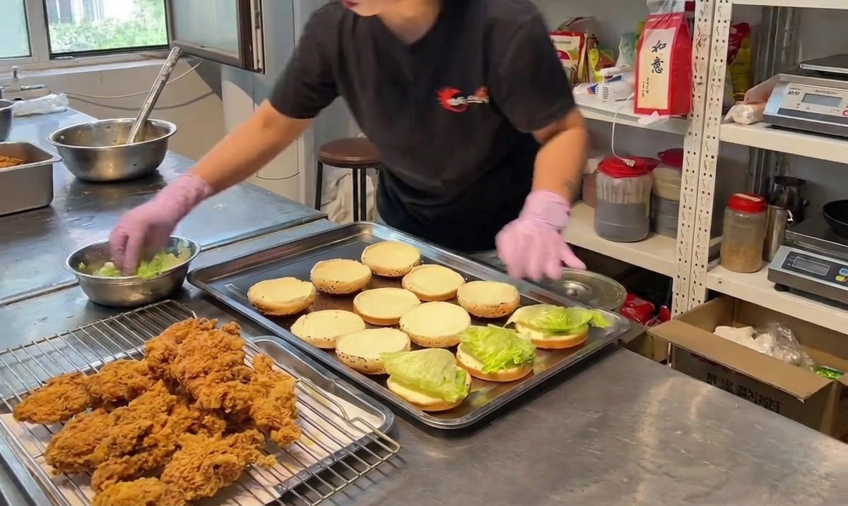 A person placing a piece of crispy fried chicken onto a bottom burger bun lined with green lettuce.