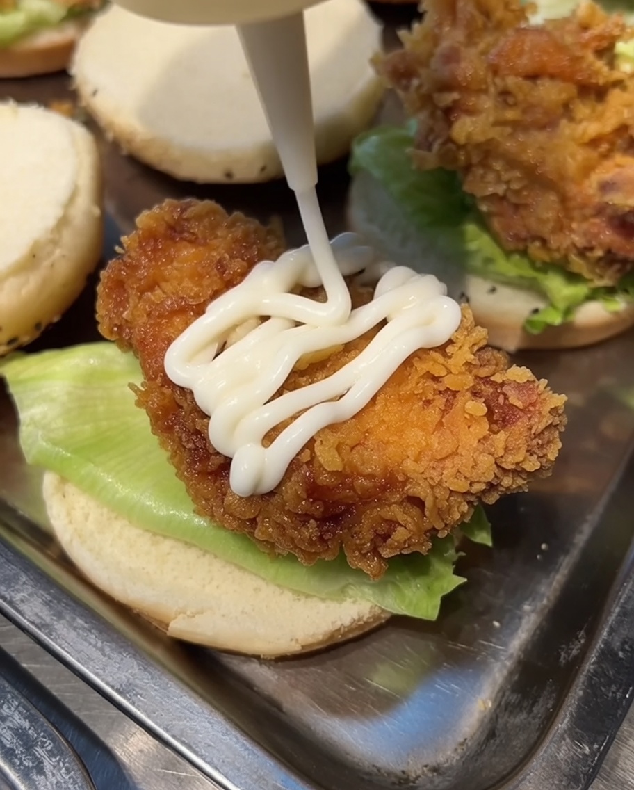 White mayonnaise being squeezed from a bottle onto a piece of fried chicken resting on a burger bun.