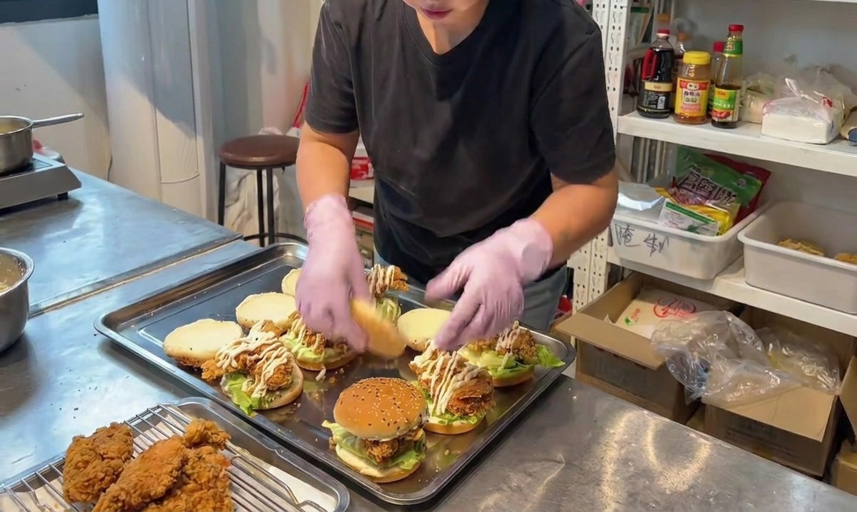 A person placing the top half of a sesame seed bun onto a fully assembled fried chicken burger.