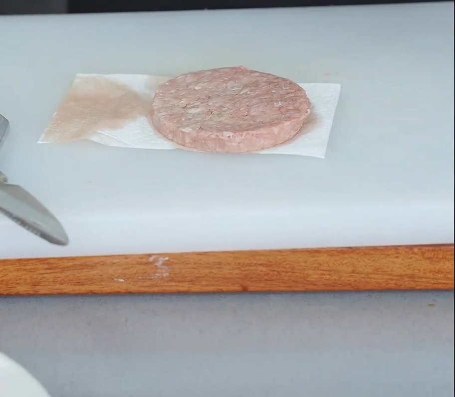 A raw beef patty resting on a paper towel with another paper towel pressed on top to absorb moisture.