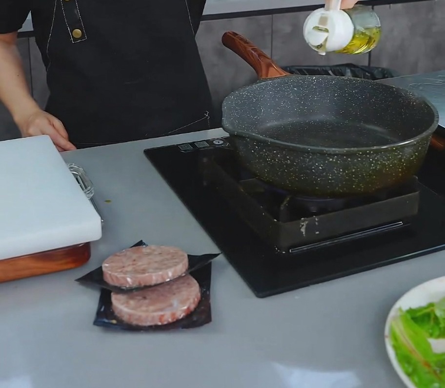 A person pouring a small amount of cooking oil from a cruet into a dark non-stick pan on the stove.