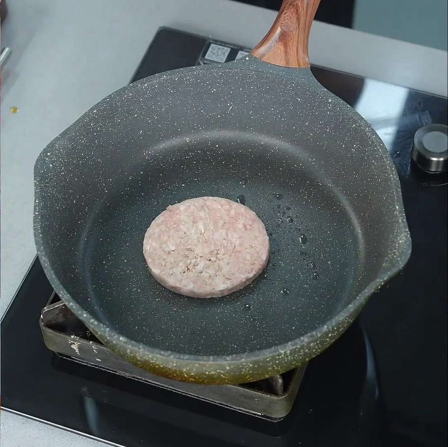 A raw beef patty beginning to sizzle in a lightly oiled pan over a stovetop burner.