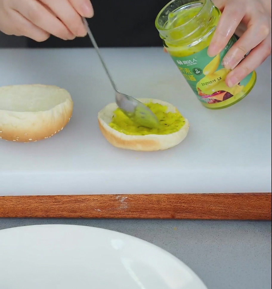 A person using a spoon to spread yellow relish onto the bottom half of a toasted hamburger bun.