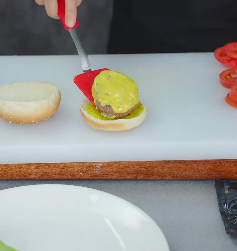 A red spatula lifting a cheese-topped beef patty onto a toasted burger bun layered with yellow relish.