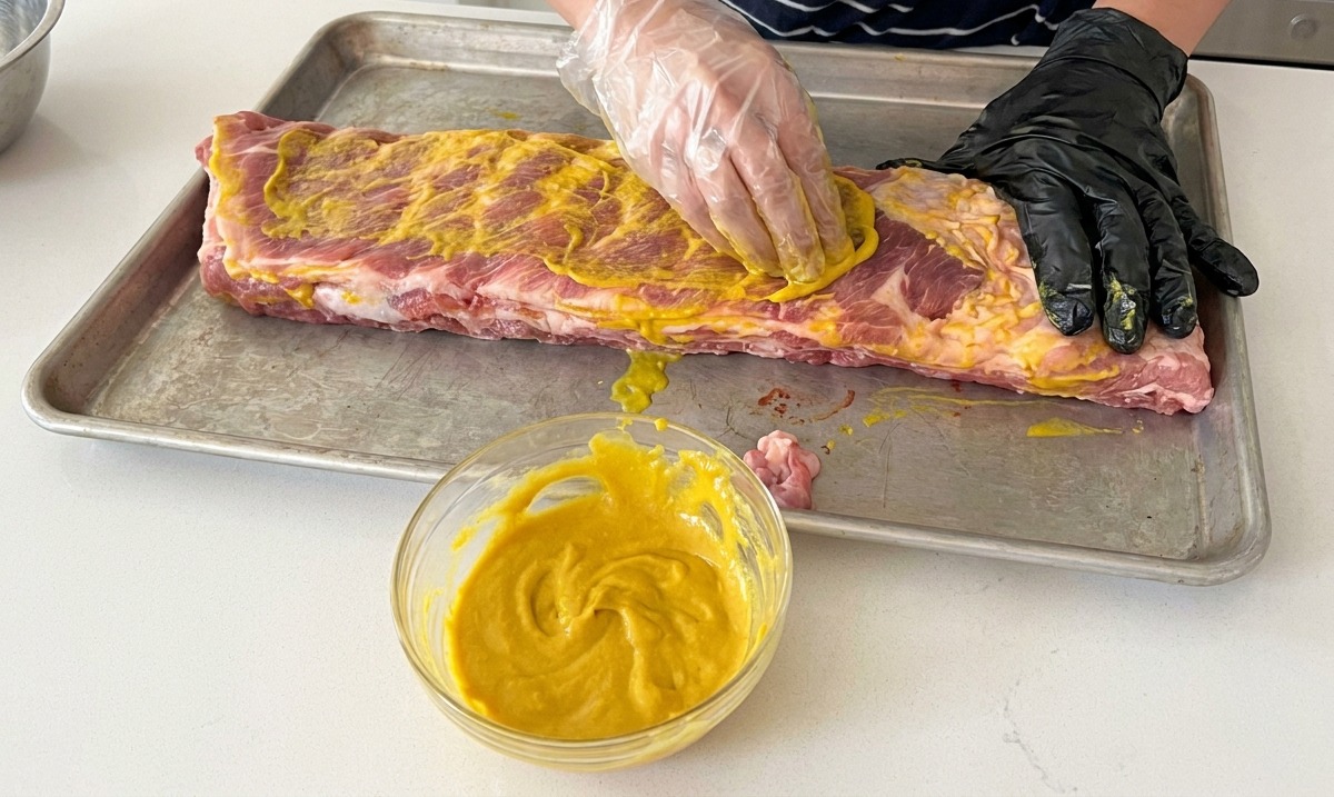 A person wearing black and clear gloves rubbing a yellow mustard binder all over raw pork ribs on a baking sheet.