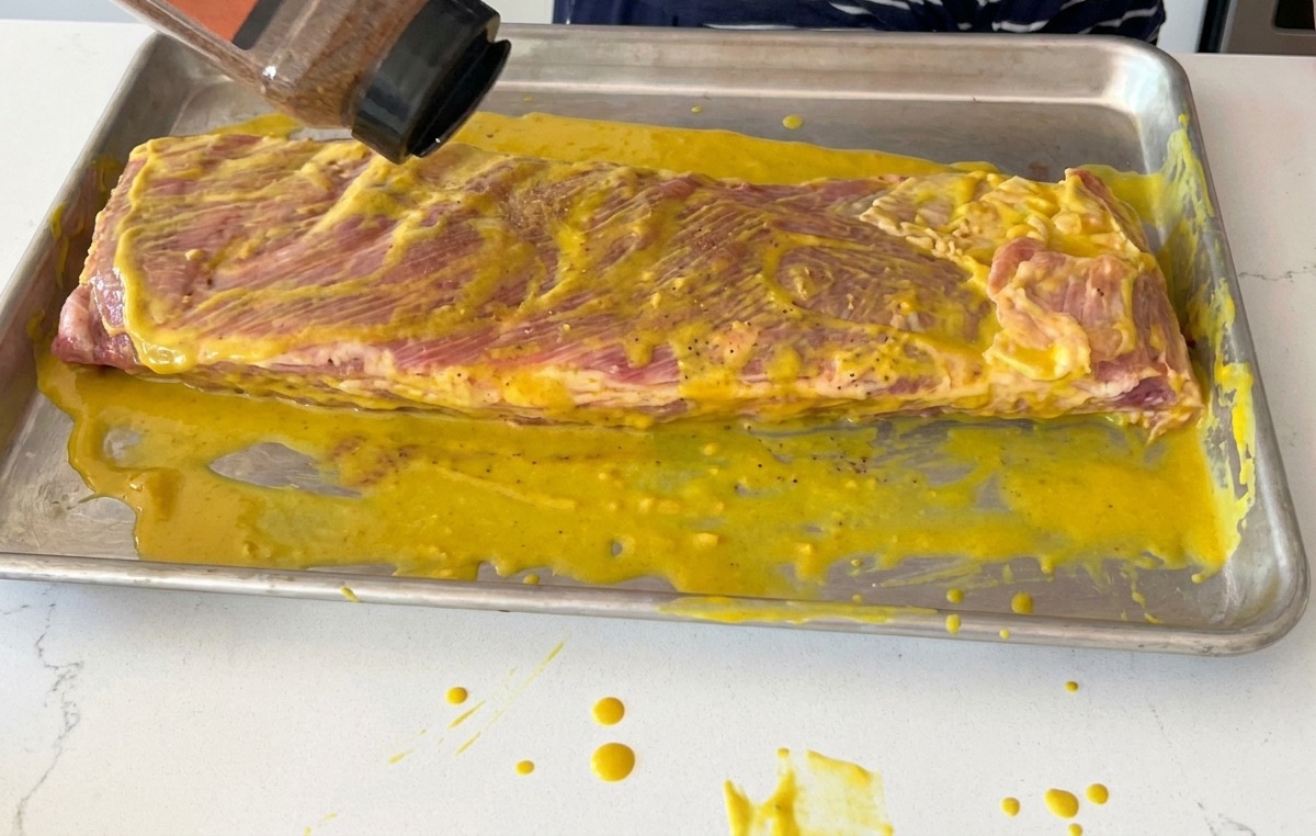 Shaking a bottle of dry BBQ rub over a rack of pork ribs coated in yellow mustard.