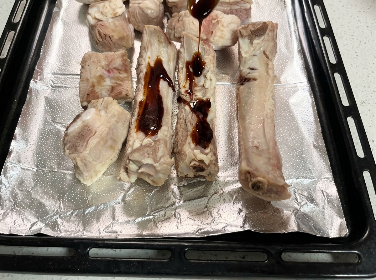Dark marinade sauce being spooned over raw pork ribs on an aluminum foil-lined tray.