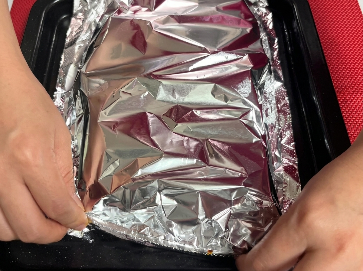 A sheet of aluminum foil being crimped and sealed over a tray of marinated pork ribs.