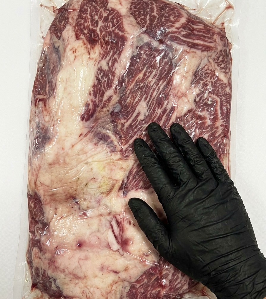 A close-up view of a knife removing thick excess fat from the surface of a raw Stone Axe Wagyu brisket.
