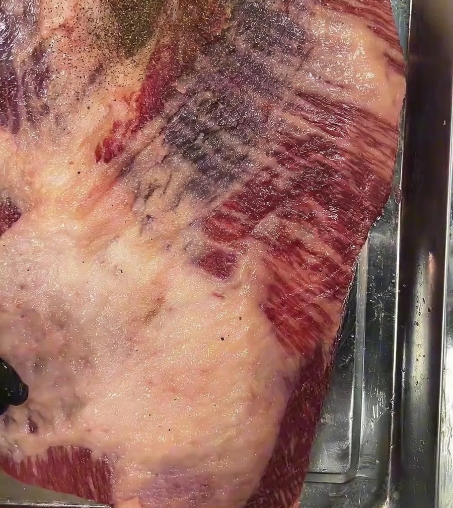 Coarse black pepper being sprinkled generously over the top of the salted raw brisket.