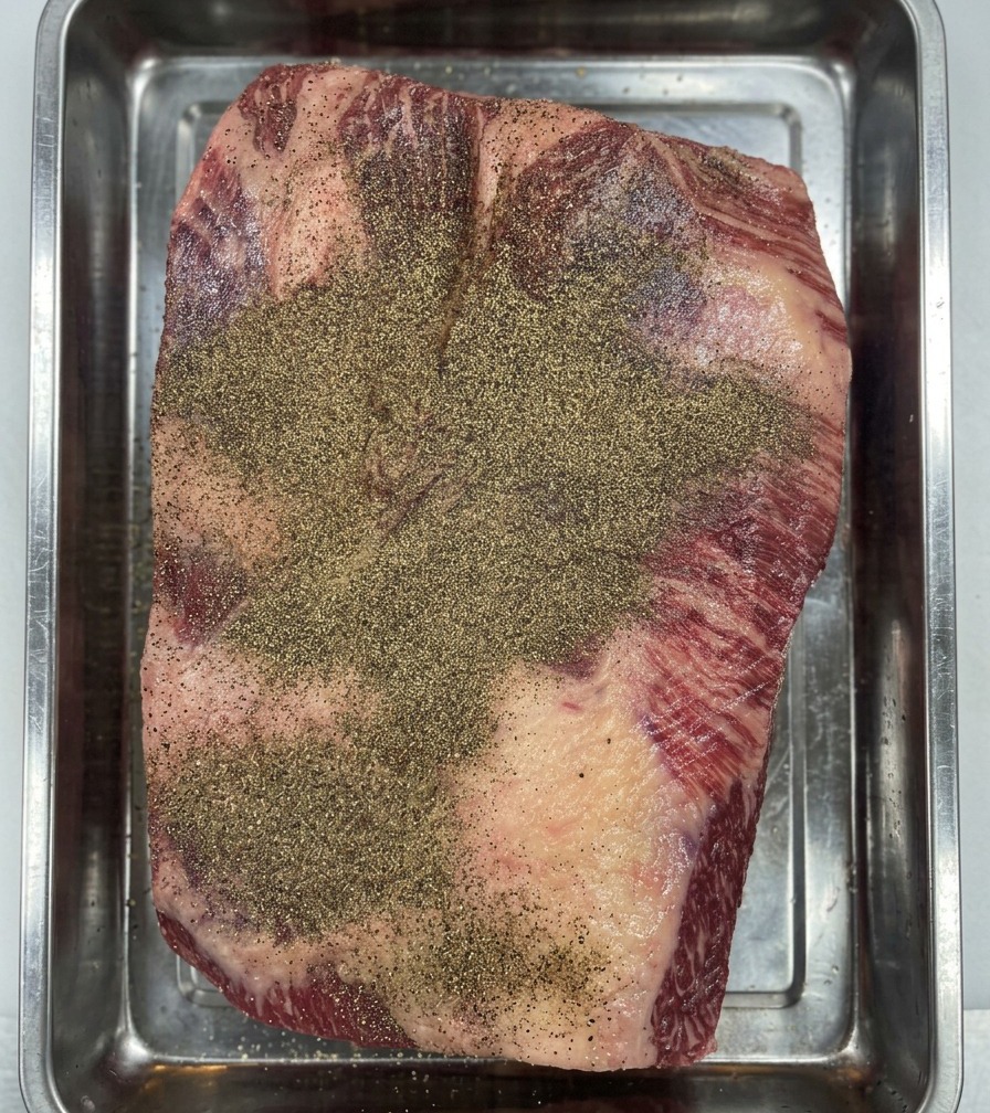 Gloved hands pressing black pepper into the surface of the Wagyu brisket to create an even seasoning layer.