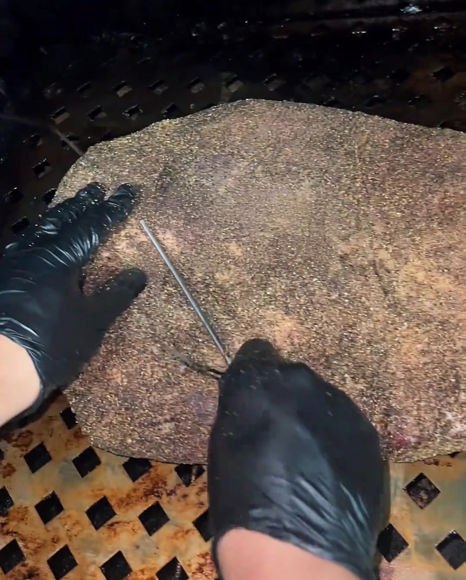 A close-up view of a thermometer probe being inserted into the thickest section of the raw, seasoned brisket.