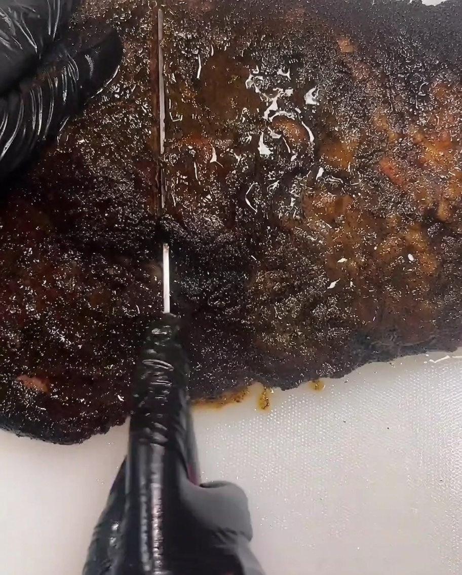 A close up of a person wearing black gloves using a long slicing knife to cut a thick dark smoked brisket on a white cutting board.