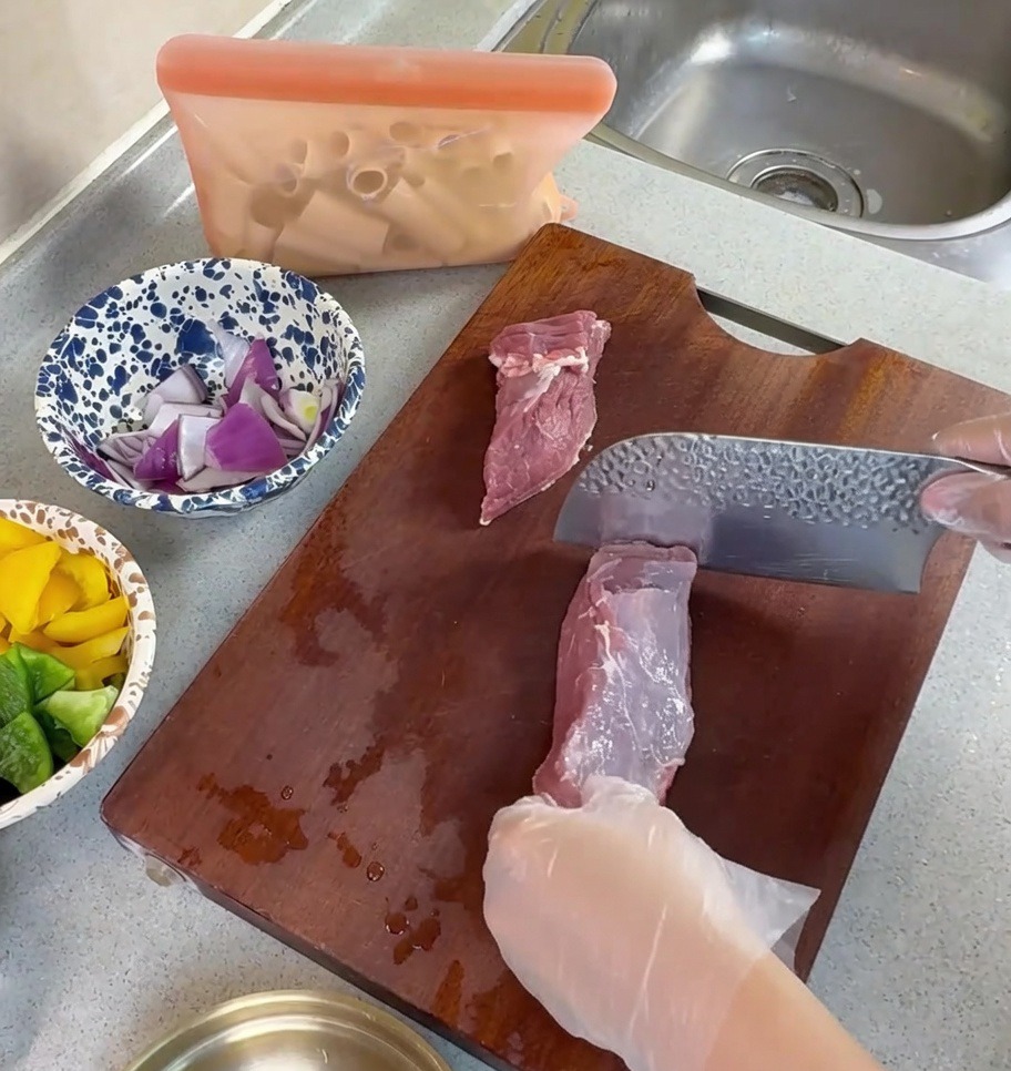 Gloved hands using a cleaver to slice raw pork into thin strips on a wooden cutting board.