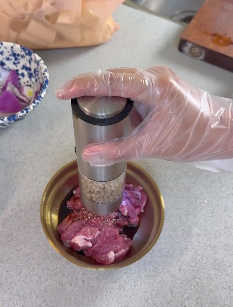 Grinding fresh pepper from an electric grinder onto sliced raw pork mixed with soy sauce in a small metal bowl.