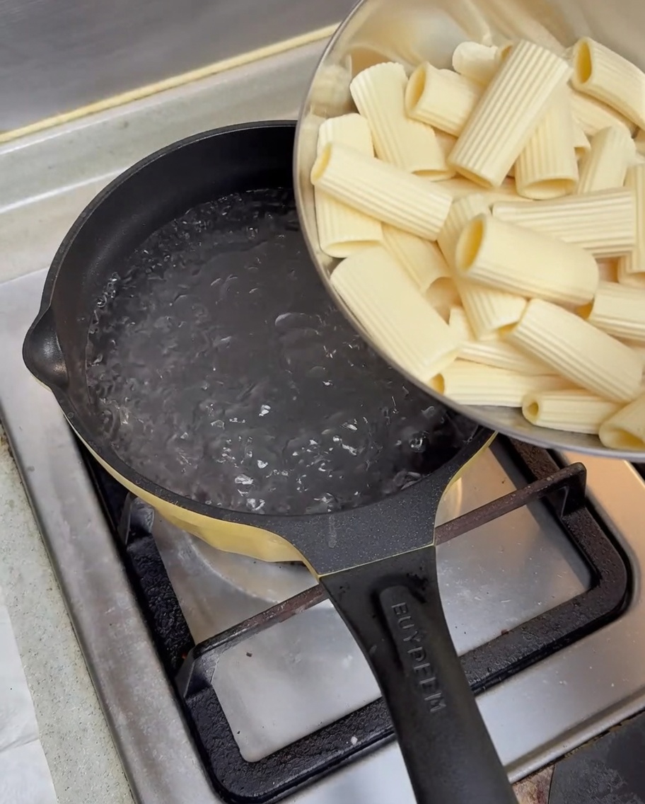 Pre-soaked rigatoni pasta being poured from a metal bowl into a pot of vigorously boiling water on a gas stove.