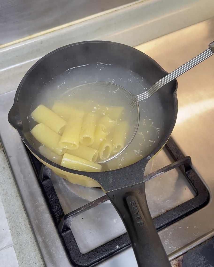 A wire skimmer lifting cooked rigatoni out of boiling water in a black pot.