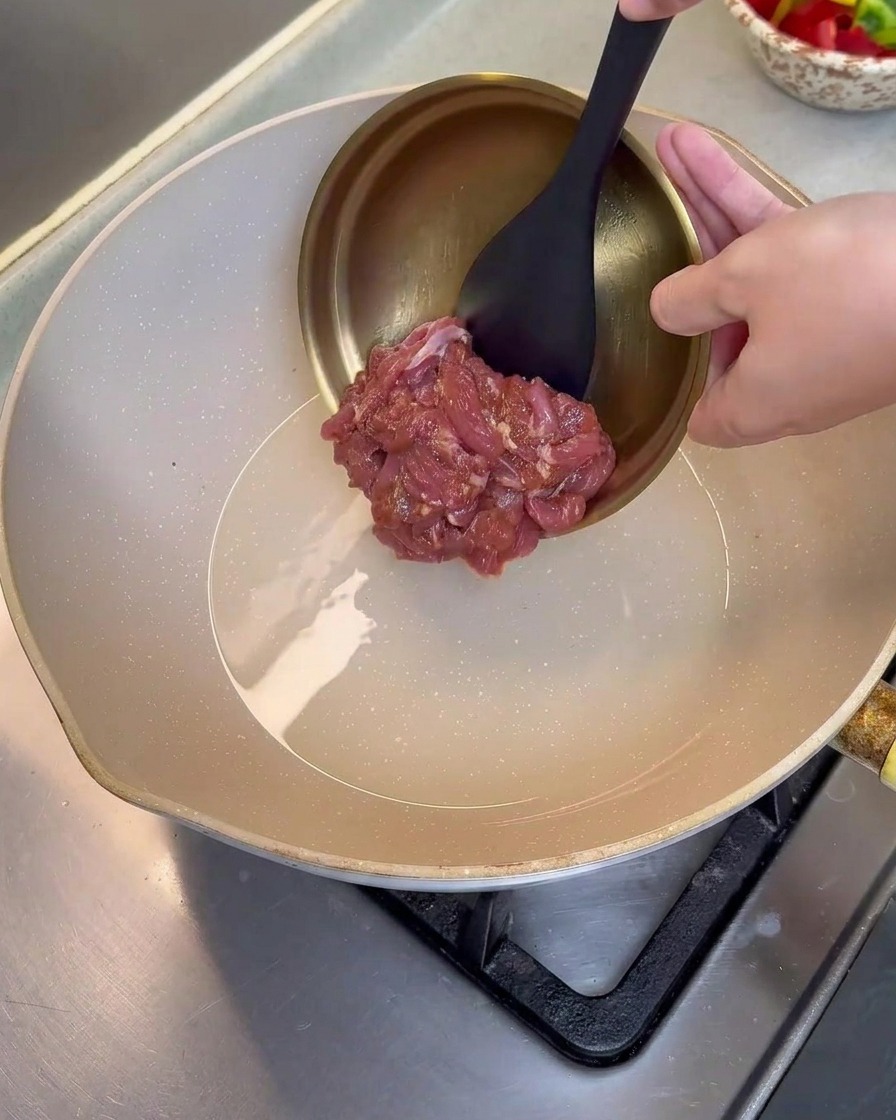 Marinated pork slices being poured from a metal bowl into a hot, lightly oiled frying pan.