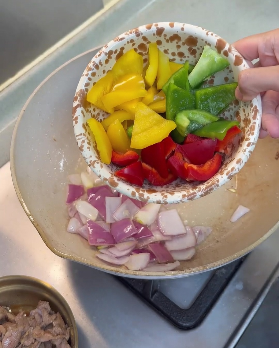 Red, yellow, and green bell pepper pieces being added to the pan with sautéed onions.