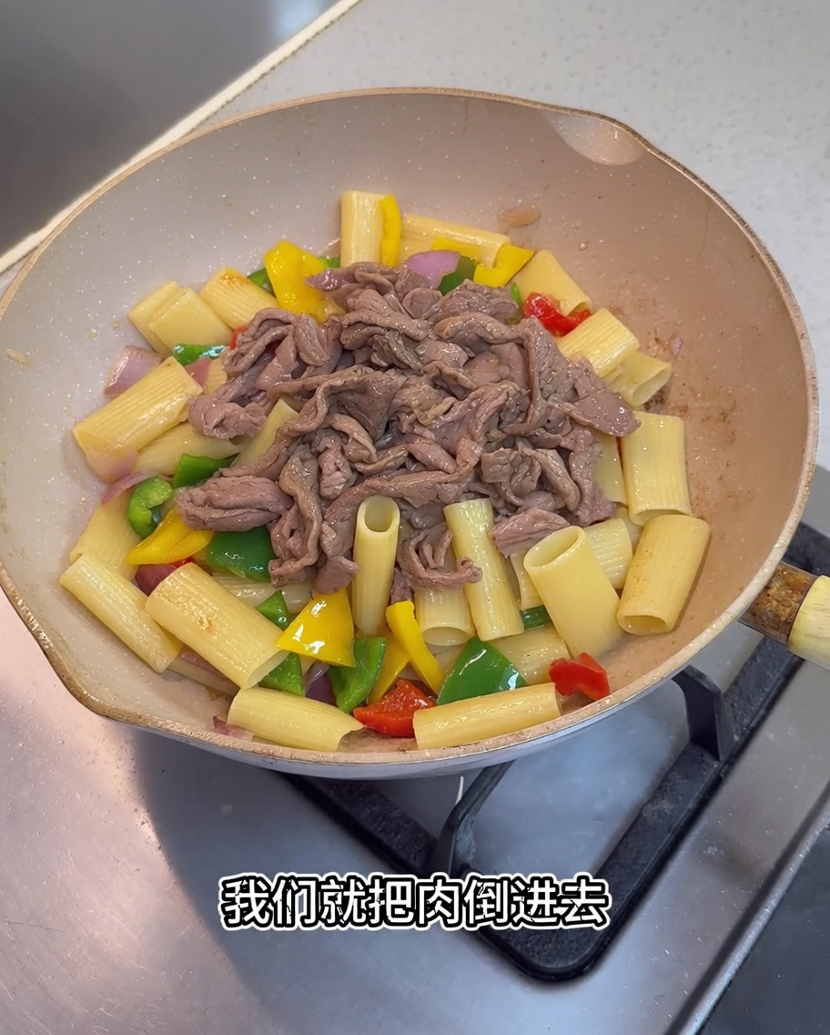 Cooked pork pieces and a dark sauce being added to the pan containing rigatoni and vegetables.
