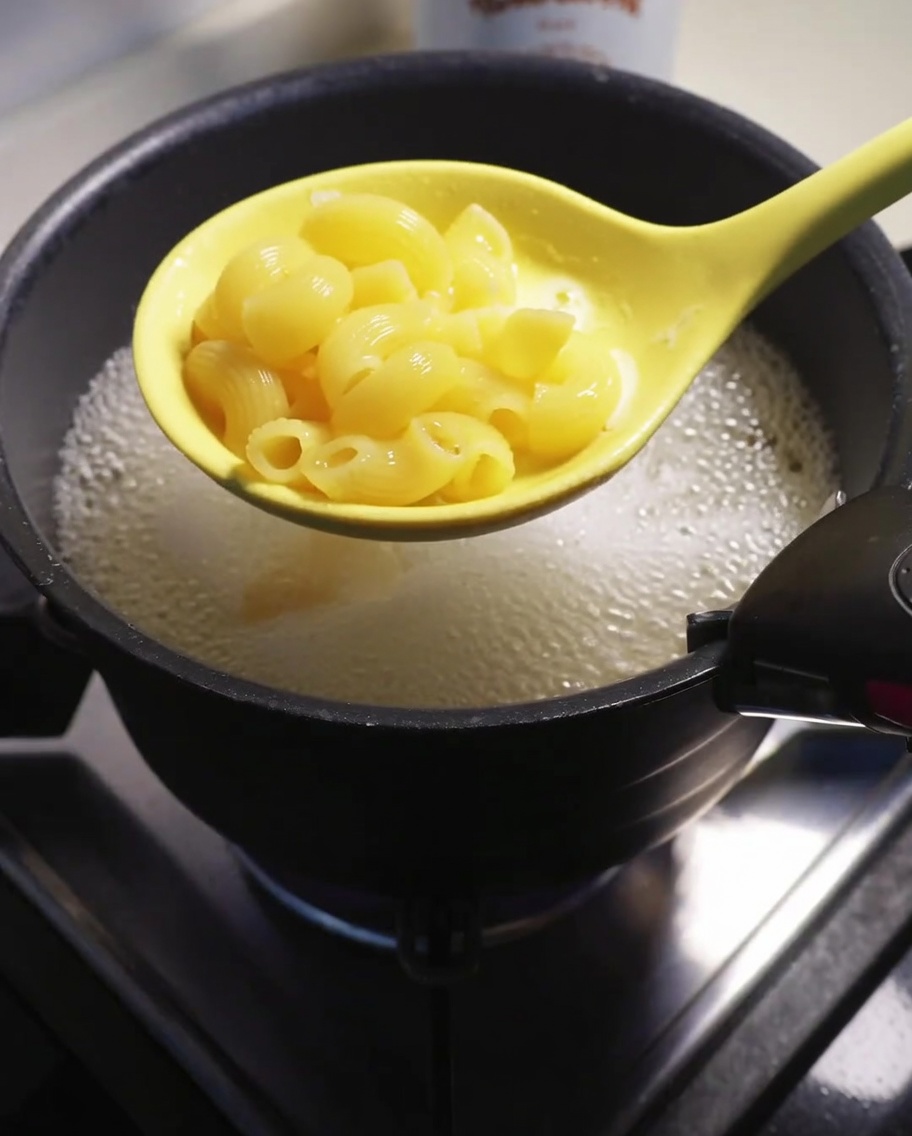 Cooked macaroni being lifted from the boiling water with a yellow slotted spoon.