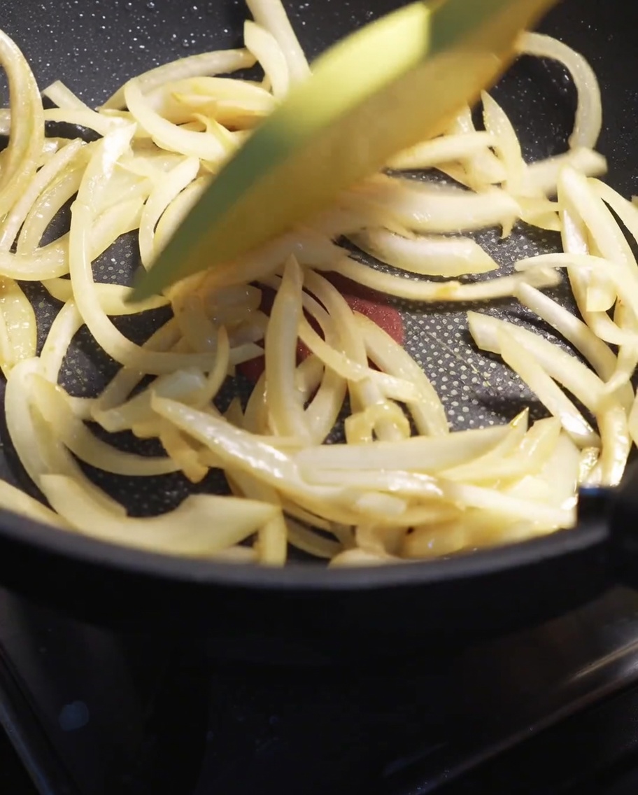 Slices of white onion being stir-fried with a spatula in a hot pan until softened.